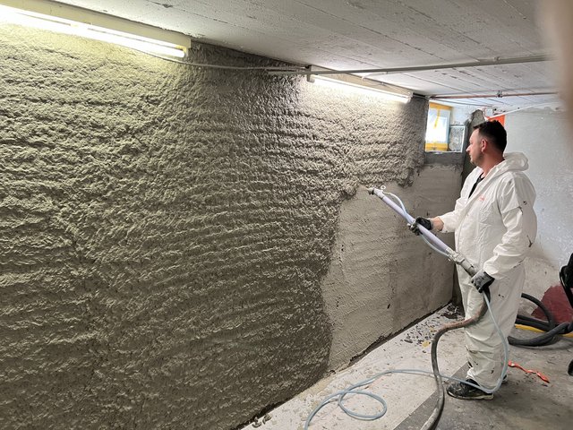 Arbeiter spritzt graue Masse auf Kellerwand auf.