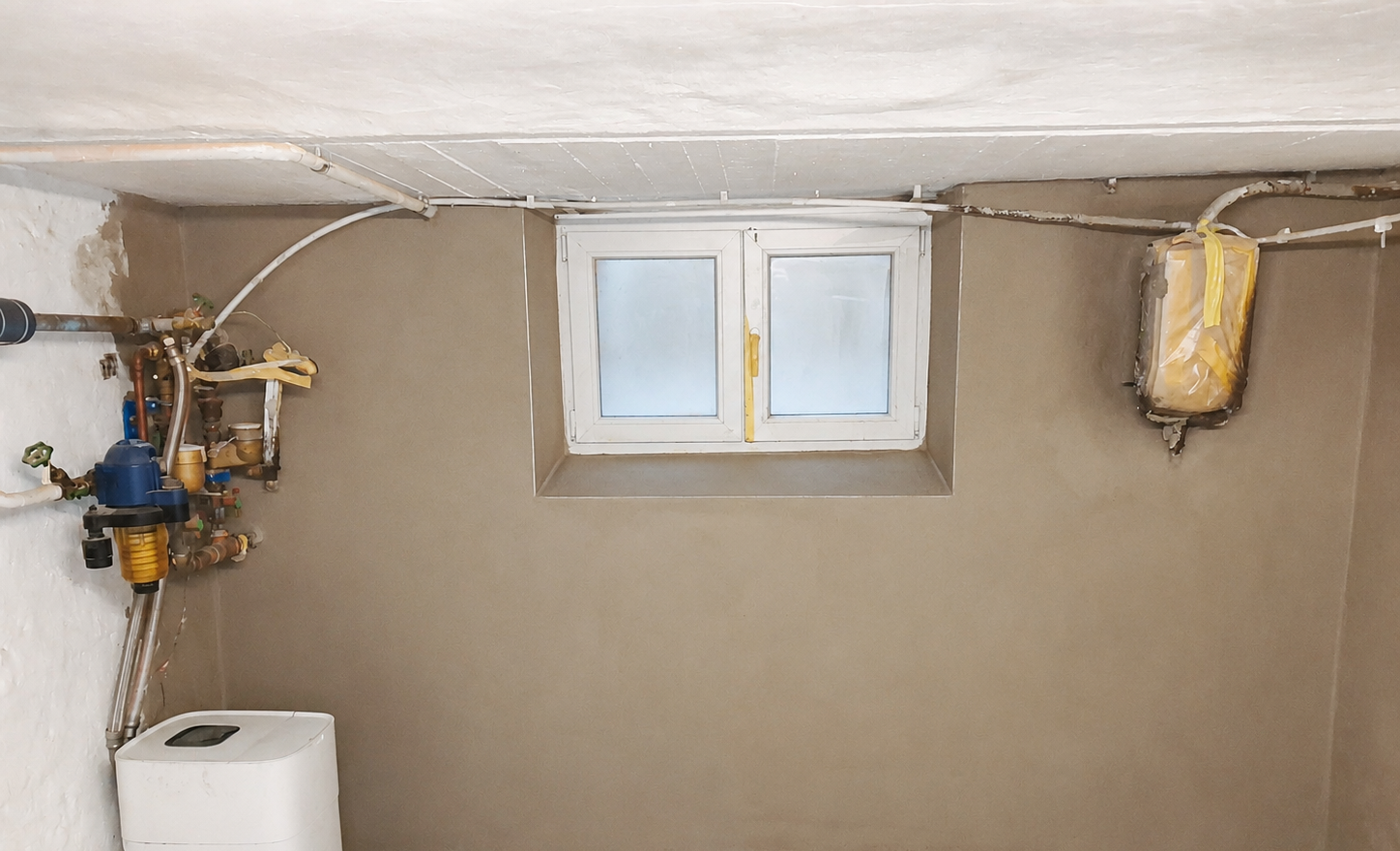 Kellerraum mit kleinem Fenster, Leitungen an der Decke und Feuchtigkeitsspuren am Wandfuß
