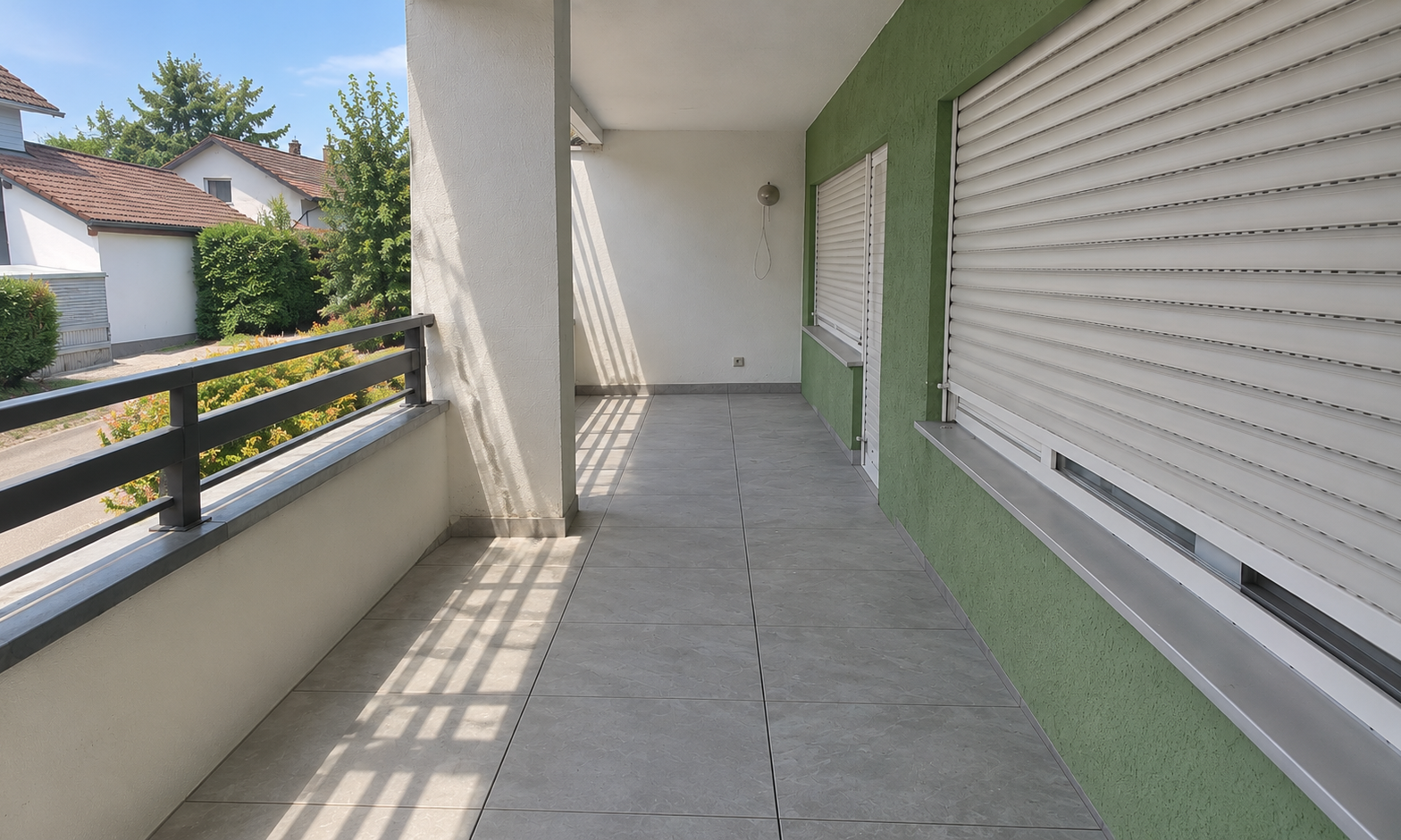 Leerer Balkon mit grauen Bodenfliesen, Metallgeländer und weißen Rollläden an grüner Wand