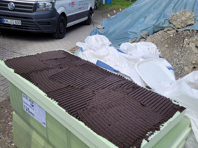 Schwarze Masse mit Zahnkelle auf heller Platte aufgetragen, Baustelle im Hintergrund