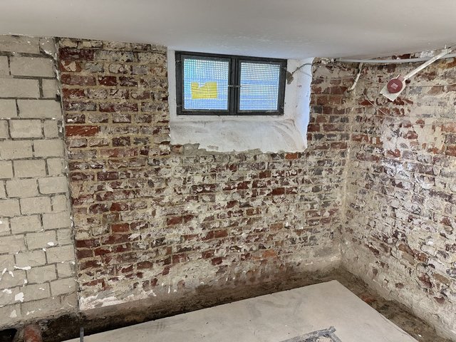 Freigelegte Ziegel-Kellerwand mit kleinem Fenster, Kabeln und Resten weißer Beschichtung