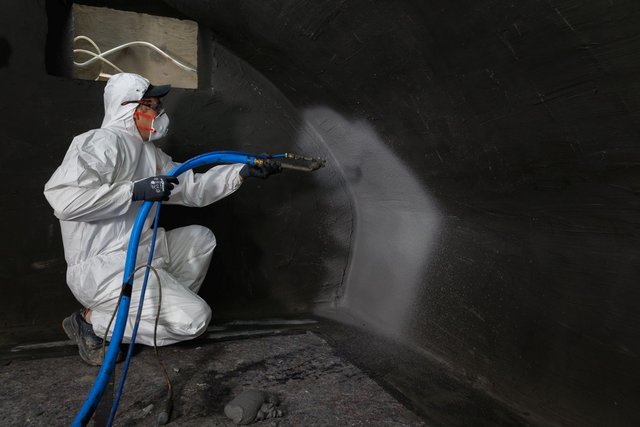 Arbeiter sprüht graue Masse auf schwarze Kellerwand, Schutzanzug und Atemmaske.
