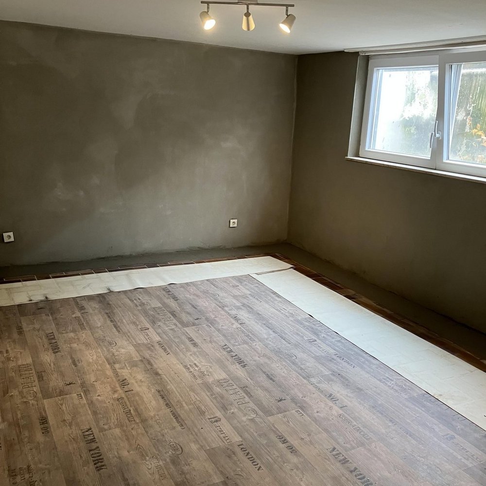 Leerer Raum mit grauen Wänden, Fenster und Holzoptikboden mit weißen Abdeckplatten am Rand
