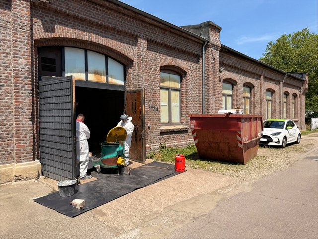 Zwei Arbeiter in Schutzanzügen bedienen eine Maschine vor Backsteingebäude.
