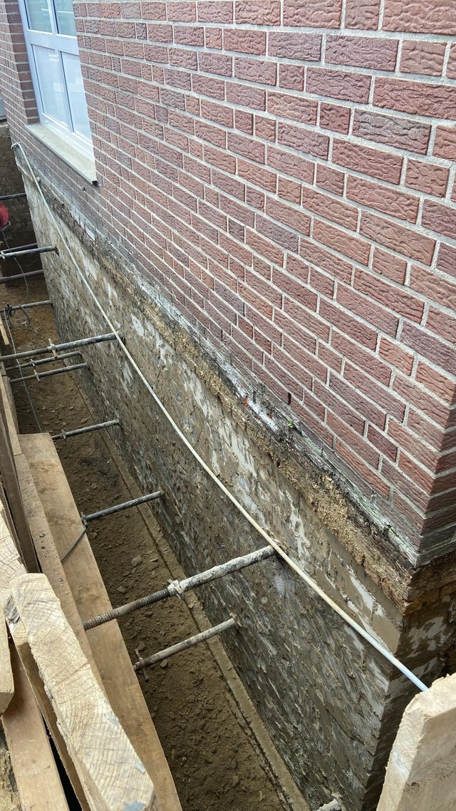 Freigelegte Kellerwand im Baugraben neben Klinkerfassade, mit Metallankern und Kabel.