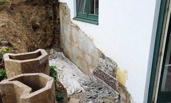 Freigelegter Gebäudesockel neben Fenster, Erdgraben und gestapelte Betonsteine im Vordergrund