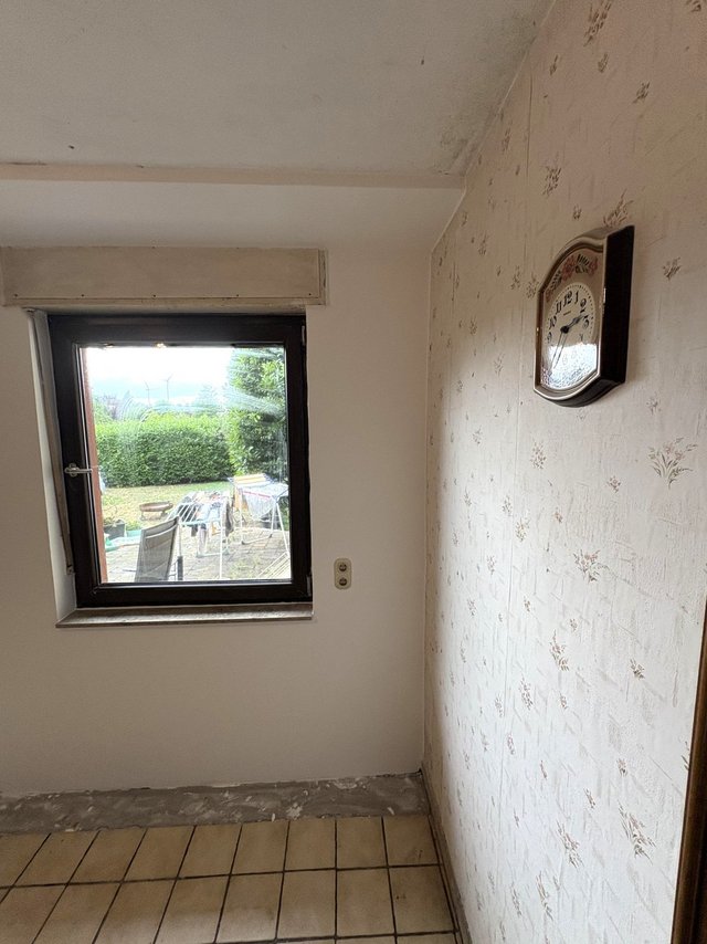 Leerer Raum mit Fenster, gemusterter Tapete, Wanduhr und Fliesenboden.
