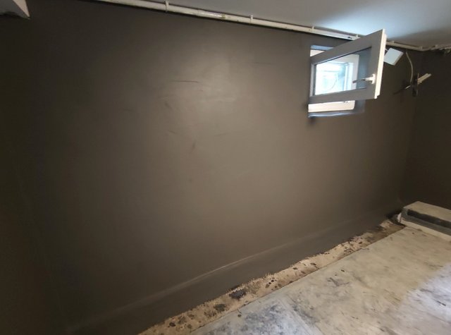 Graue Wandfläche mit kleinem geöffnetem Kellerfenster und Rohren an der Decke