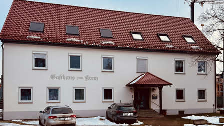 Weißes Gebäude mit rotem Ziegeldach, Schriftzug „Gasthaus zum Kreuz“ und zwei Autos davor.