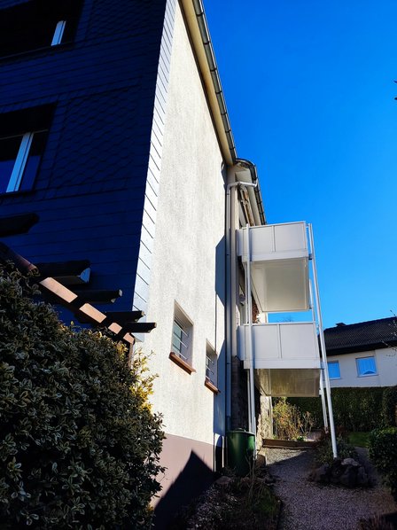 Hausfassade mit Balkonen und Regenrinne unter blauem Himmel, Perspektive von unten.