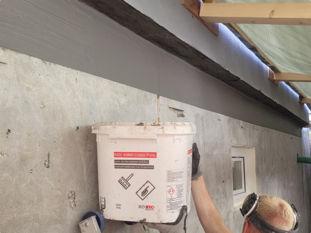 Arbeiter trägt schwarze Masse auf Betonwand unter Holzdecke auf.
