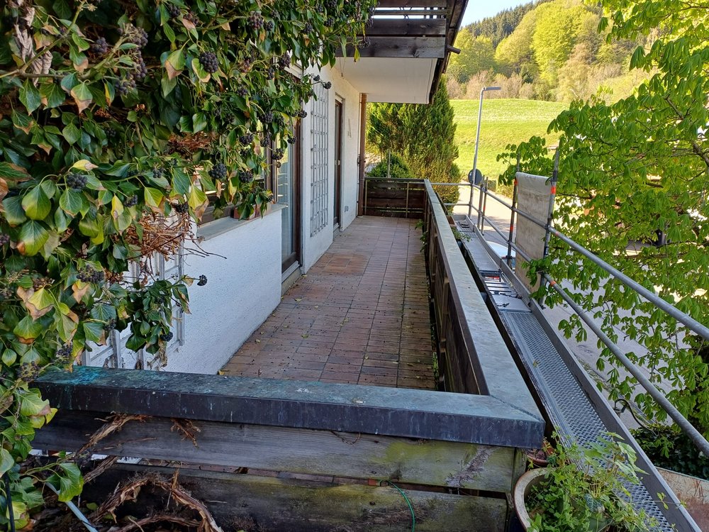 Balkon mit Fliesenboden, Holzgeländer und Gerüst neben Haus, umgeben von Grün.