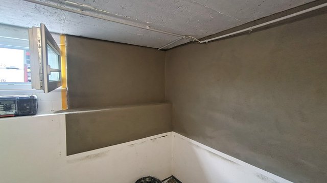 Graue Masse auf Kellerwand, Fenster geöffnet, Leitungen an Betondecke sichtbar.