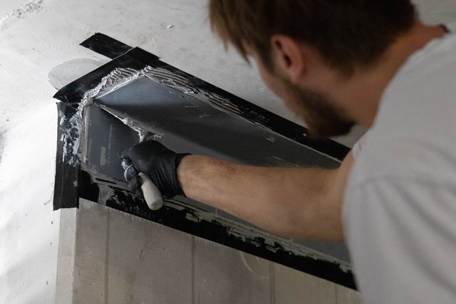 Arbeiter trägt graue Masse mit Spachtel auf Wandfläche neben abgeklemmter Folie auf.