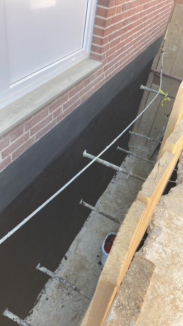 Schwarze Beschichtung an freigelegter Hauswand im Arbeitsgraben neben Fenster.