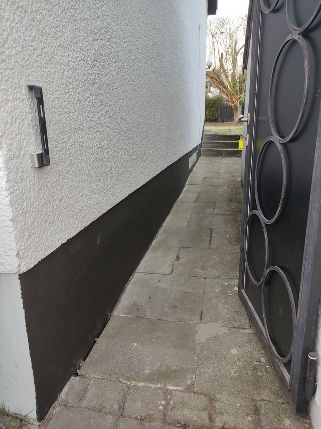 Schmaler Steinplattenweg neben Hauswand mit schwarzem Sockelanstrich und geöffnetem Metalltor