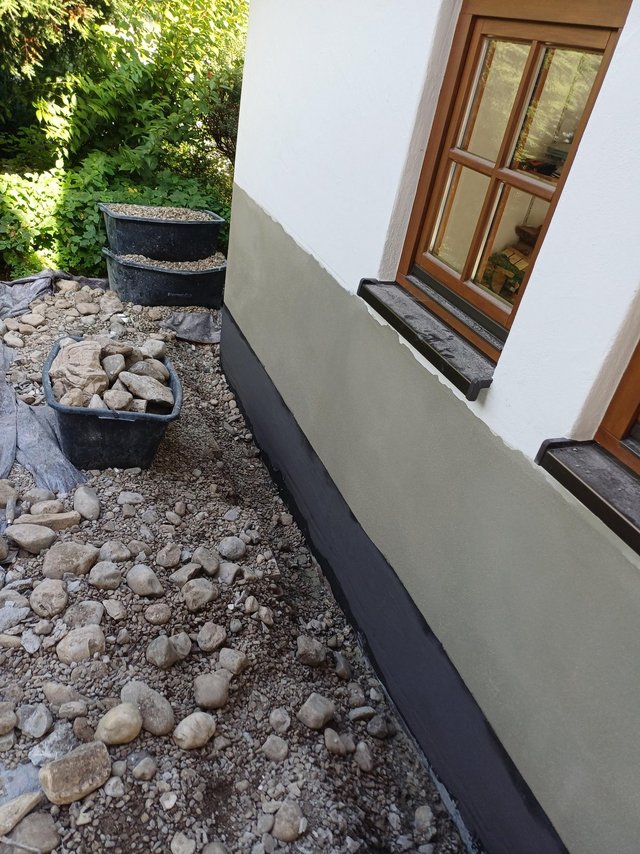 Hauswand mit schwarzer und grauer Beschichtung, davor Kies und Steinkübel