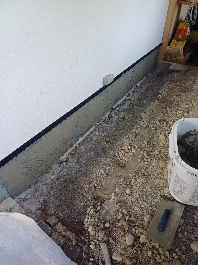 Graue Masse auf Sockelbereich einer Hauswand, Baustelle mit Schotterboden
