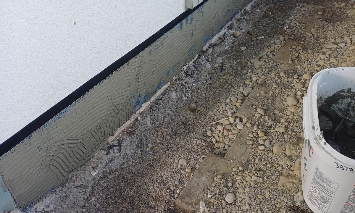 Graue Masse auf Sockelbereich einer Hauswand, Baustelle mit Schotterboden