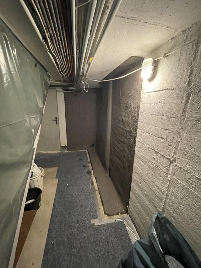 Schmaler Kellerflur mit sichtbaren Leitungen und Betonwänden.