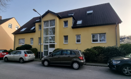 Gelbes Mehrfamilienhaus mit dunklem Satteldach, Eingang und parkenden Autos