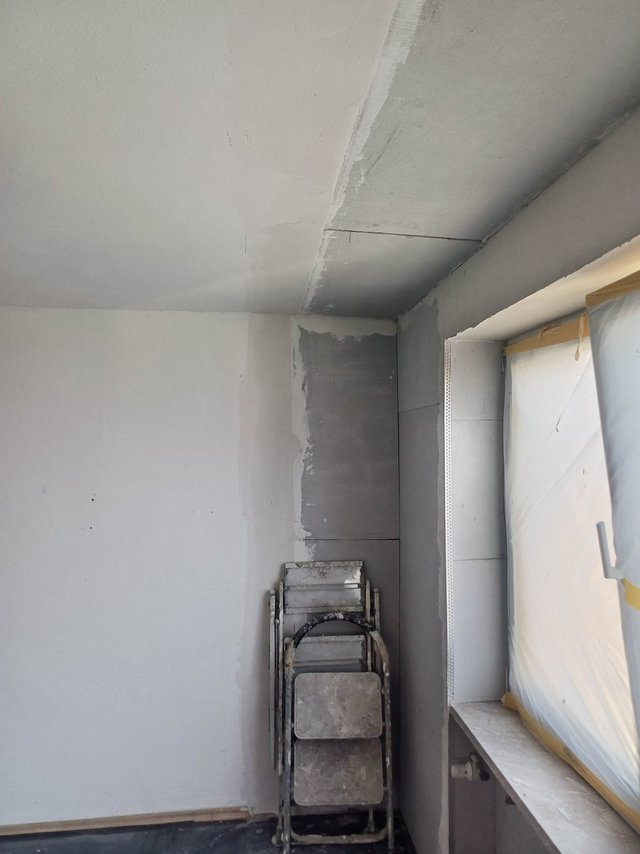 Leere Zimmerecke mit grauen Platten, davor Klapptritt, Fenster mit Folie abgedeckt
