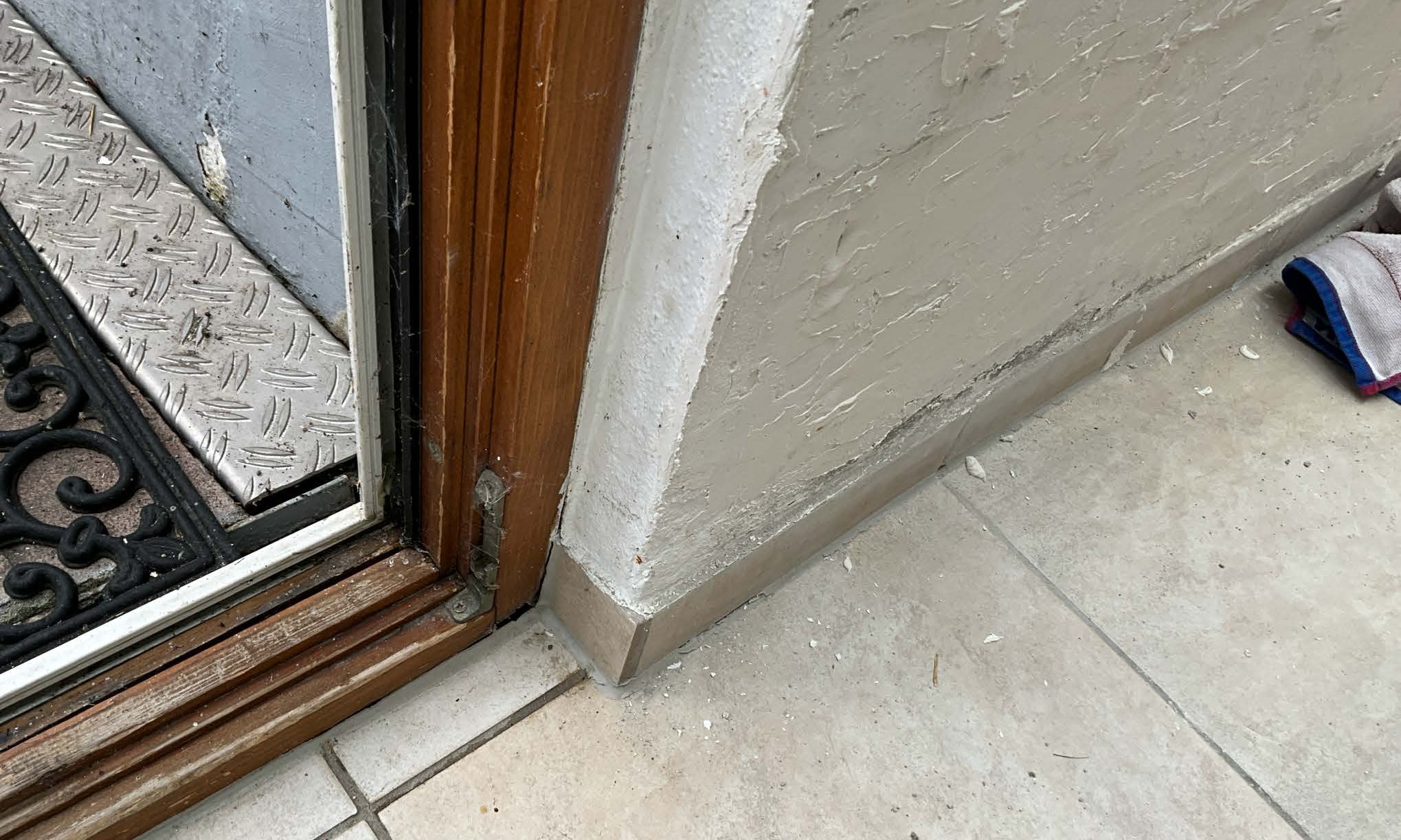 Abgeplatzter Wandputz am Sockel neben Holzrahmen und Fliesenboden.
