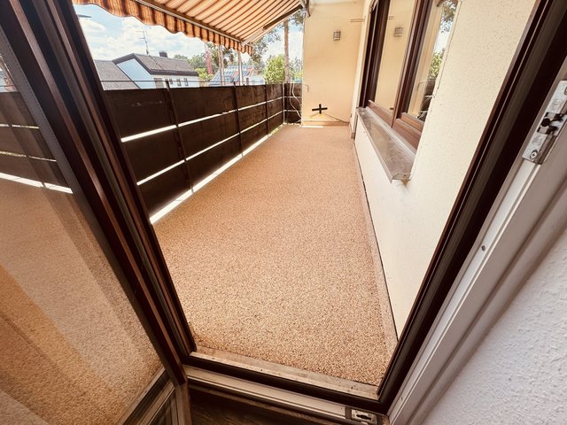 Blick durch geoeffnete Balkontuer auf Balkon mit Markise, dunkler Verkleidung und Steinbelag