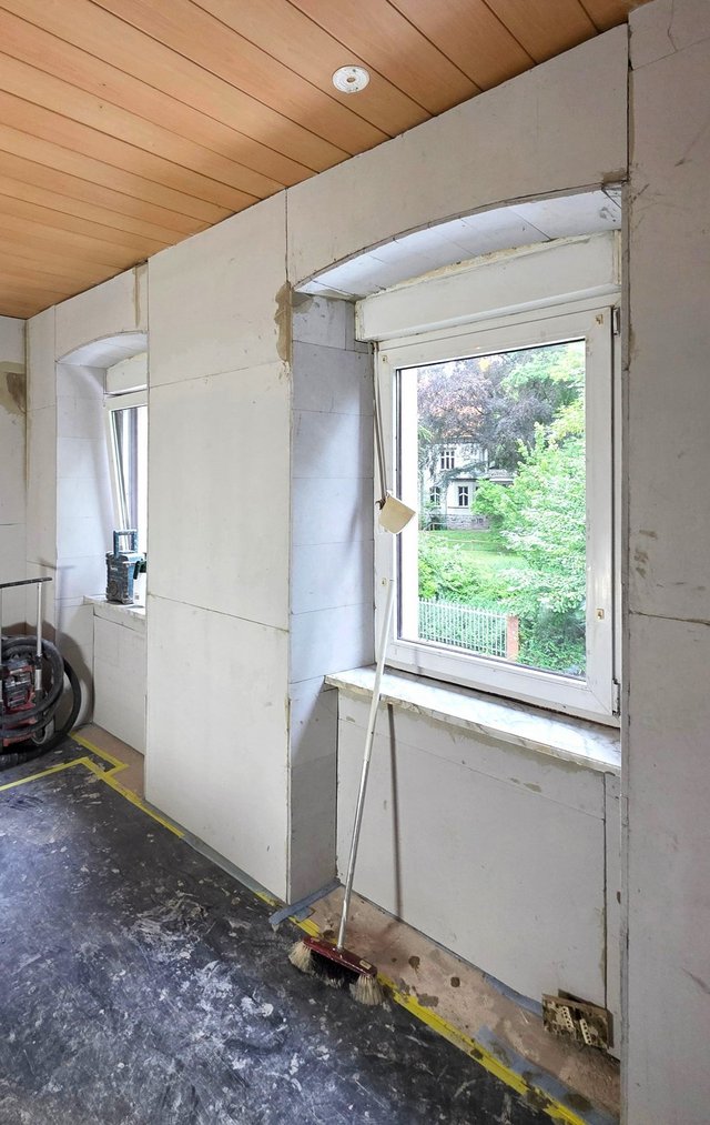Baustellenraum mit Fenster, helle Platten an Wand und Holzdecke, Besen lehnt am Sims.