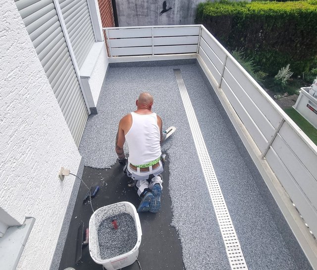 Arbeiter verteilt graue Steinchen auf Balkonboden neben Entwaesserungsrinne.