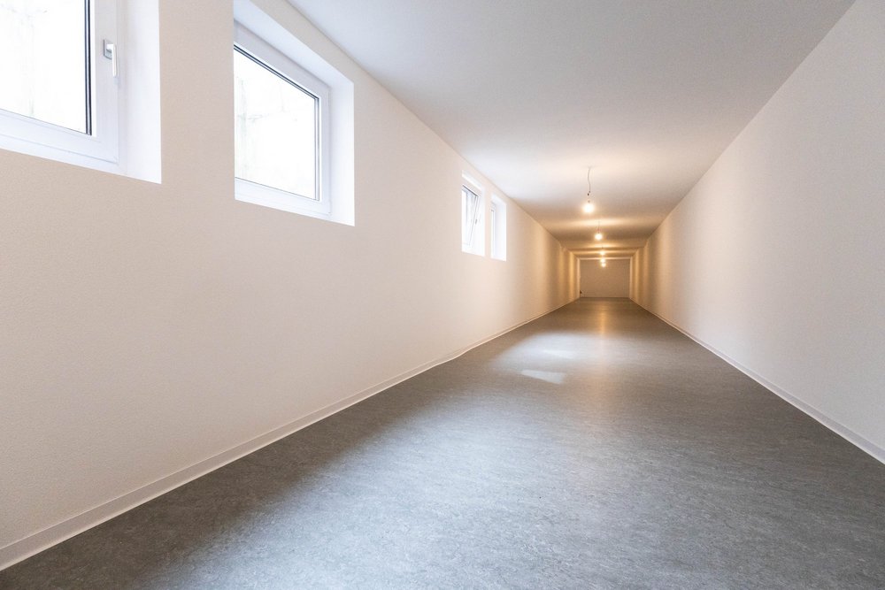 Langer leerer Kellerraum mit grauem Boden, weißen Wänden und kleinen Fenstern