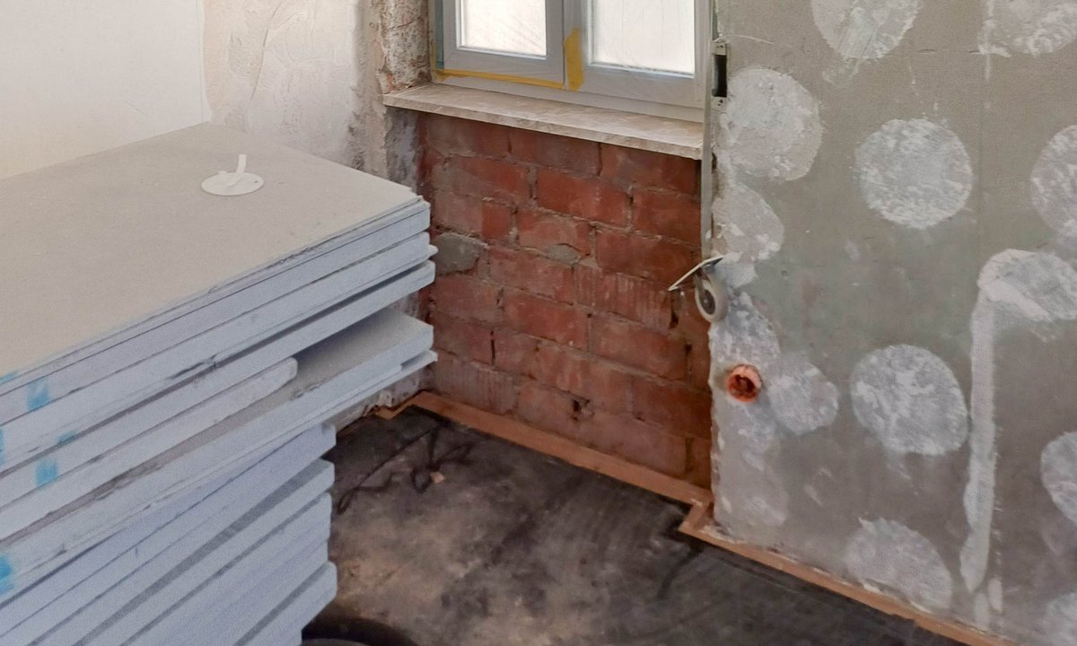 Baustelle: Fenster mit Schutzfolie, Wand mit grauen Masse-Tupfen, freigelegtes rotes Ziegelmauerwerk
