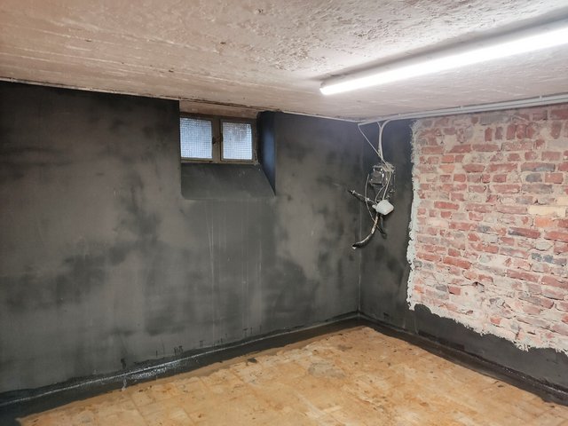 Kellerraum mit schwarz beschichteter Wand, freigelegter Ziegelwand und kleiner Fensteröffnung