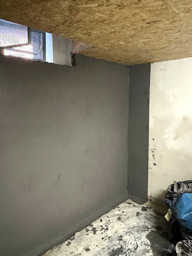 Graue Wandbeschichtung an Kellerwand unter Holzdecke, Fenster mit Gitter.