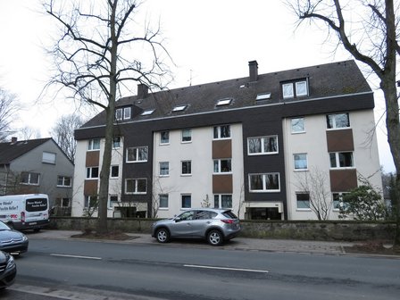 Mehrfamilienhaus an Straße mit geparkten Autos und kahlen Bäumen, Frontansicht