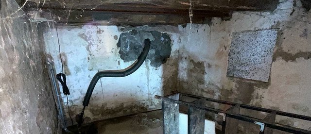 Schwarzer Schlauch führt durch Maueröffnung in feuchten Kellerraum, metallenes Gestell davor
