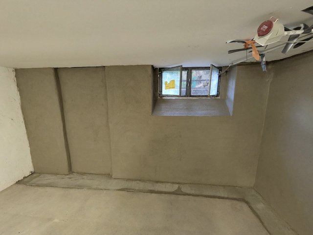 Kellerraum mit verputzten Betonwaenden und kleinem Fenster mit Metallgitter.