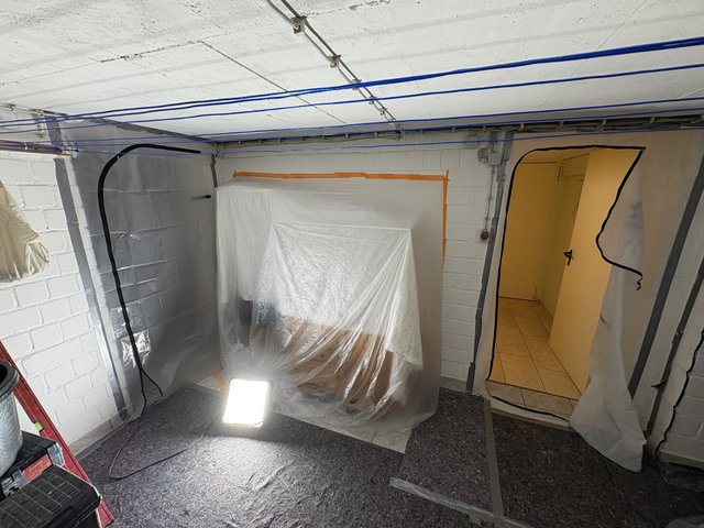 Kellerraum mit Folienabklebung, abgedeckten Gegenständen und Baustrahler am Boden