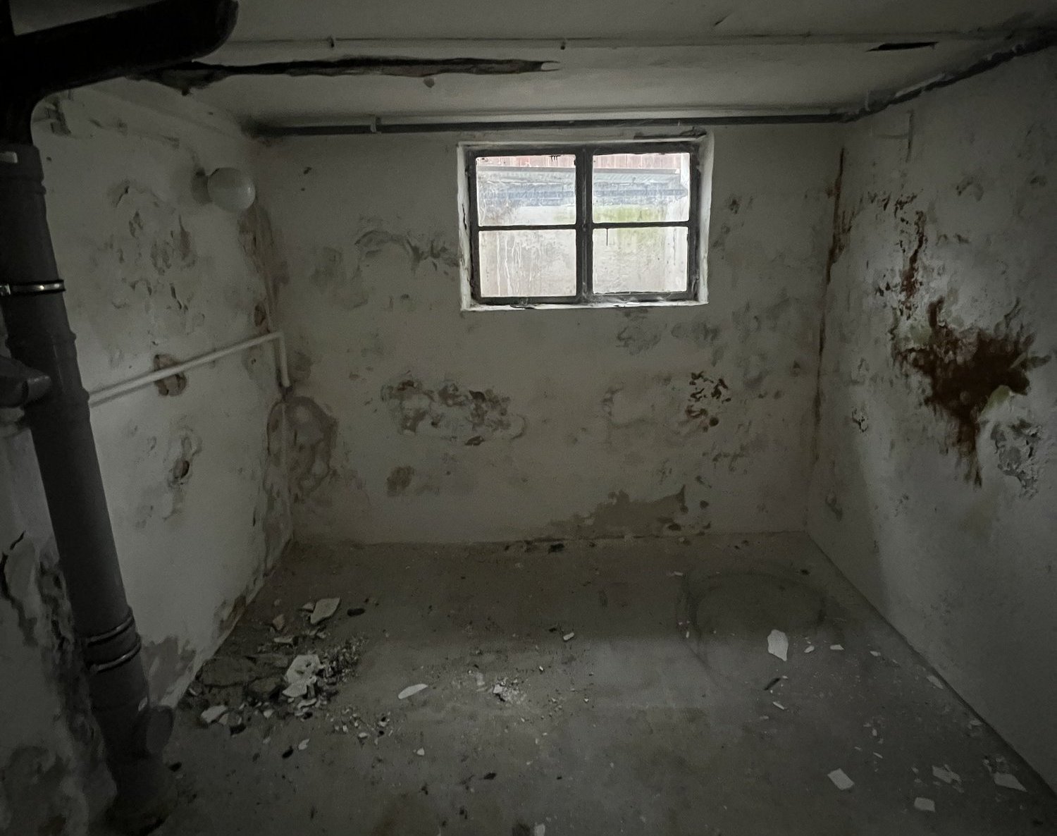 Kellerraum mit fleckiger Wand und kleinem Fenster, Schutt auf dem Boden