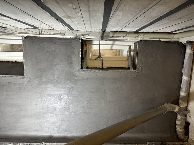 Kellerwand mit grauer Masse beschichtet, zwei kleine Fenster und freiliegende Leitungen