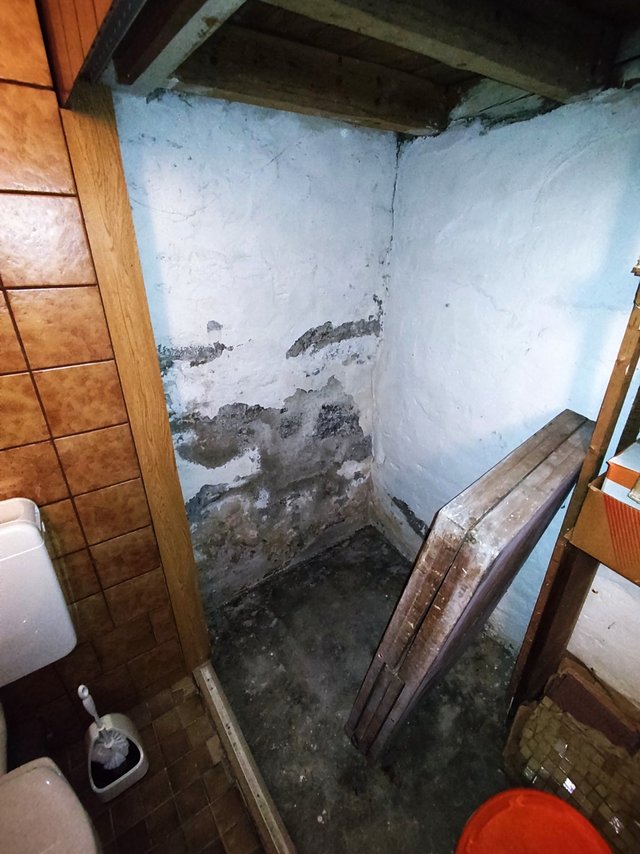 Fleckige, beschädigte Kellerwand in Abstellnische neben gefliester Toilettenecke.