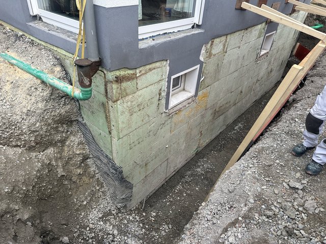 Freigelegte Kellerwand mit grünen Dämmplatten und Lichtschächten im Erdgraben