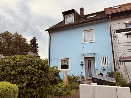 Hellblaues Wohnhaus mit Geruest am Anbau und bewachsenem Vorgarten