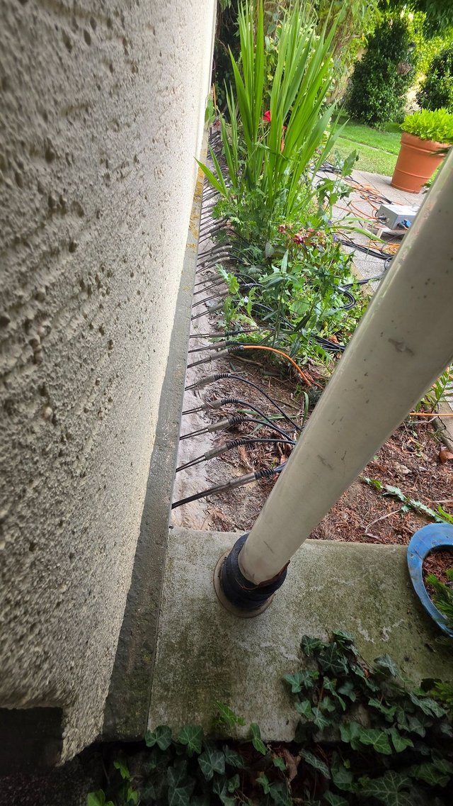 Mehrere Schläuche und Kabel verlaufen entlang einer Betonwand im Gartenbereich.