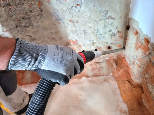 Handschuhhand saugt Bohrlochstaub aus Lochreihe in Ziegelmauer.