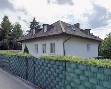 Wohnhaus mit dunklem Dach hinter grünem Sichtschutzzaun.