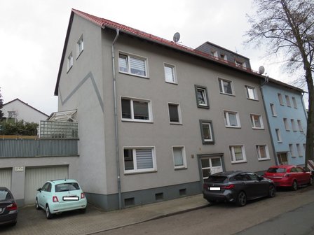 Mehrstöckiges Wohnhaus mit grauer Fassade, roten Dachziegeln und parkenden Autos an der Straße.