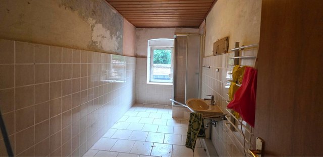 Schmaler, gefliester Badraum mit Dusche, Waschbecken und abblätternden Wandflächen
