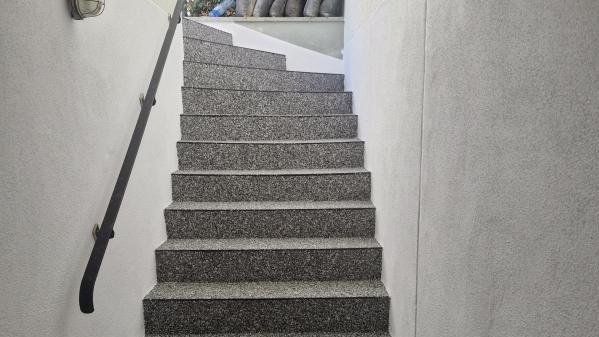 Graue Steintreppe im Treppenhaus mit Handlauf und Wandlampe, Blick nach oben.
