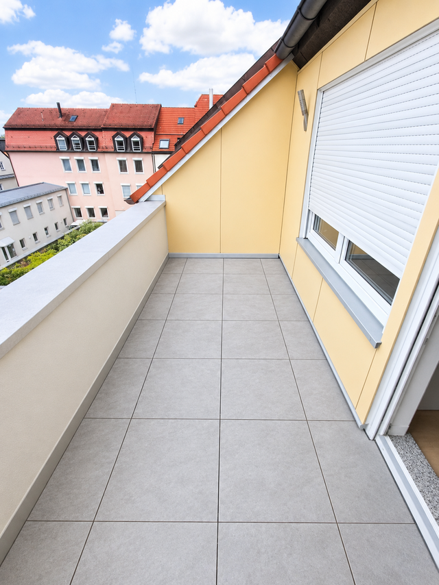Schmaler Balkon mit grauen Bodenfliesen, gelber Wand und geschlossenem Rollladen